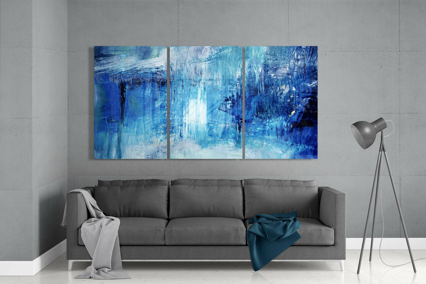 Three-panel abstract blue glass wall art hung above gray sofa in minimalist interior with floor lamp.