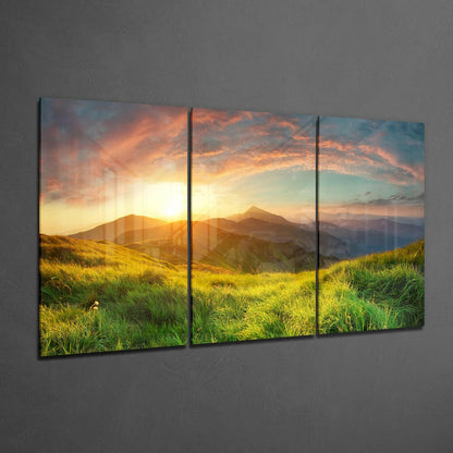 Three-panel glass wall art displaying a mountain sunset over grassy hillside, mounted on gray textured wall.