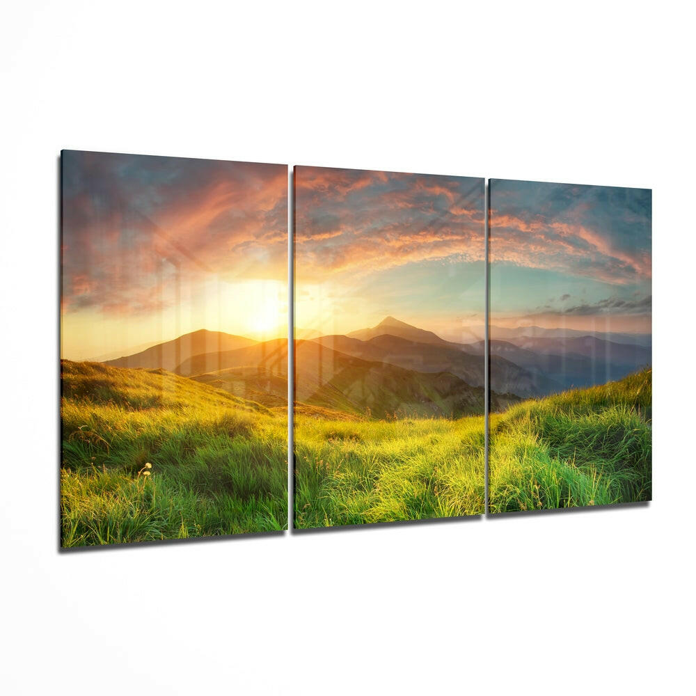 Three-panel glass wall art displaying a golden sunset over green hillsides and distant mountains.