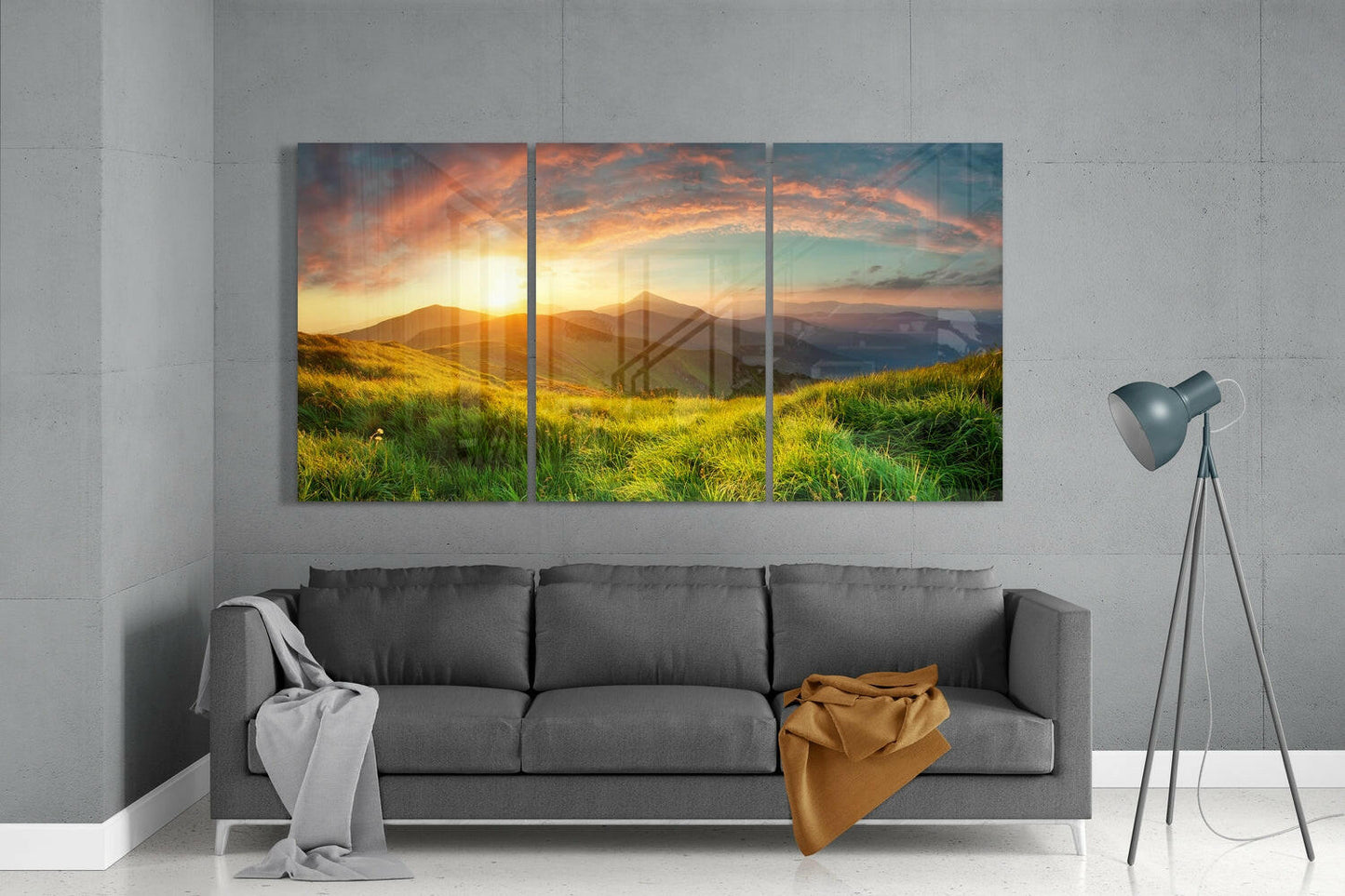Three-panel glass wall art depicting a golden sunset over rolling green hills and distant mountains, displayed above a gray sofa in a modern living room.