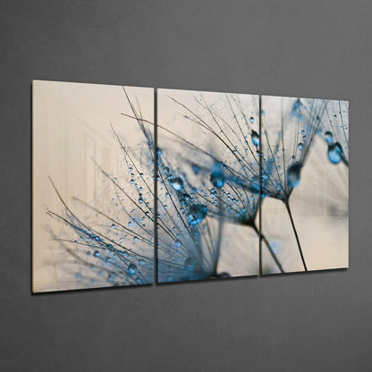 Three-panel glass wall art displaying a close-up dandelion with blue-tinted water droplets against a cream background, mounted on gray wall.