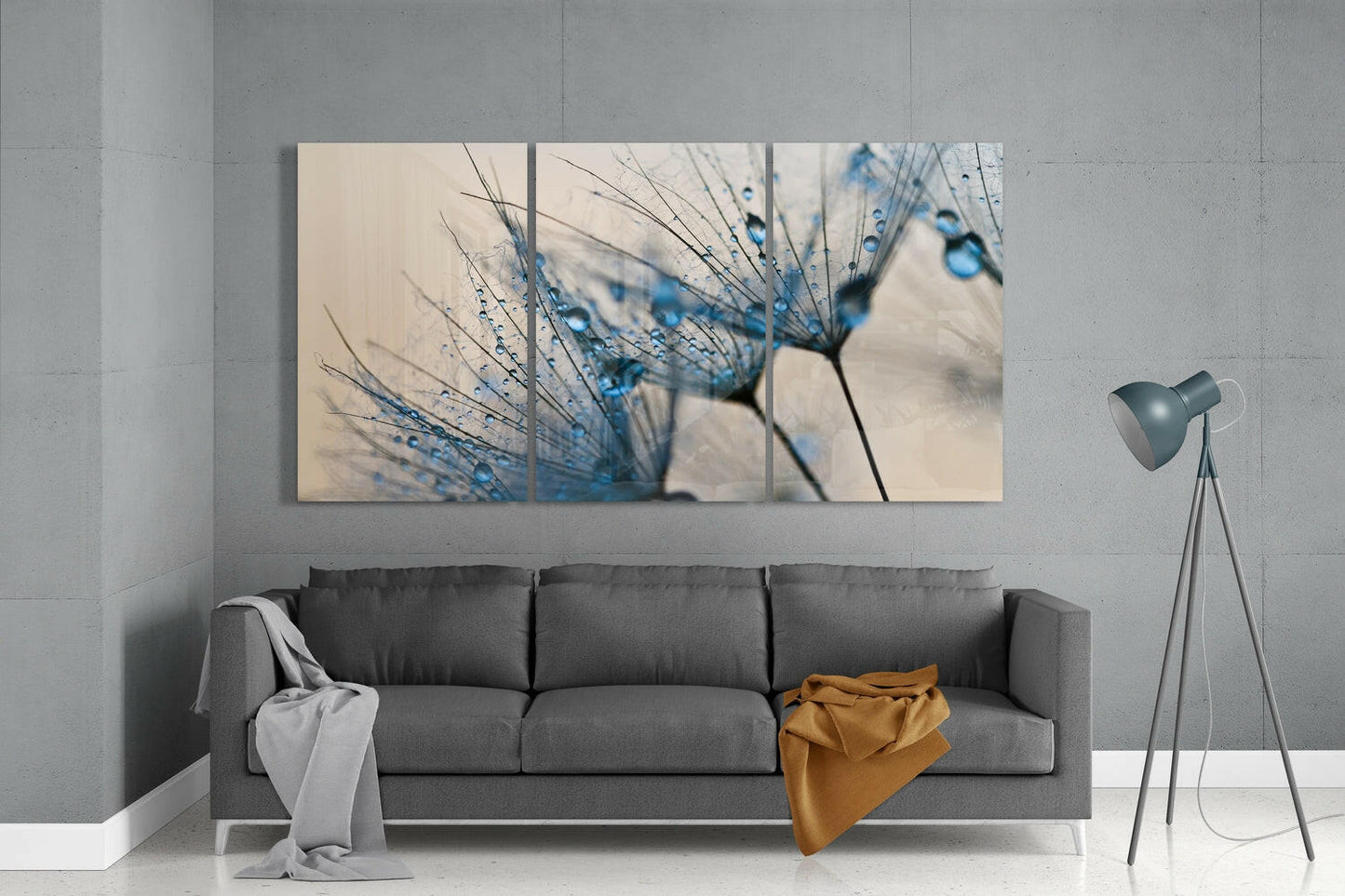 Three-panel glass wall art above gray sofa displaying blue water droplets on dandelion seeds against cream background.