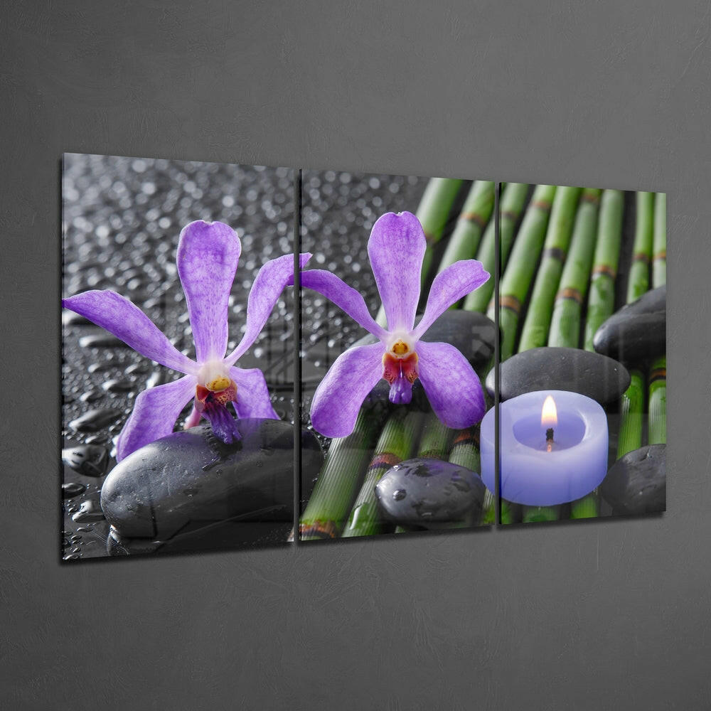 Three-panel glass wall art displaying purple orchids with black stones, green bamboo, and a lit lavender candle against gray wall.