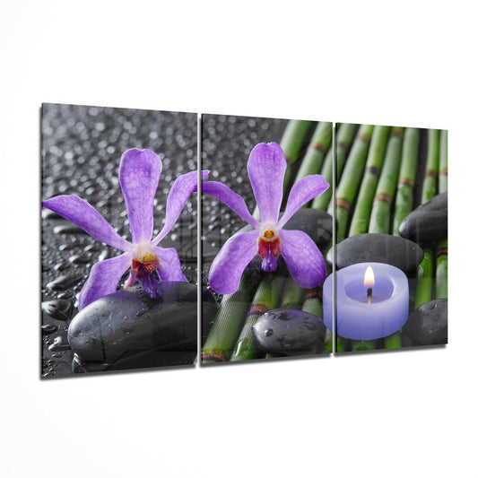 Three-panel glass wall art displaying purple orchids with dark stones, green bamboo, and a lit purple candle against blurred bokeh background.
