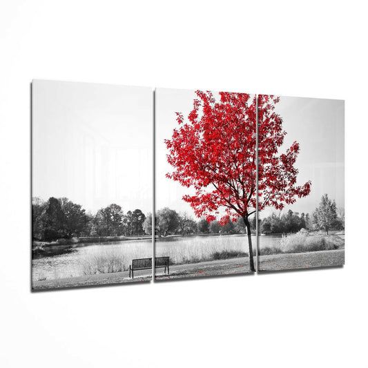 Three-panel glass wall art displaying a black and white landscape with a vibrant red tree beside a bench overlooking water.