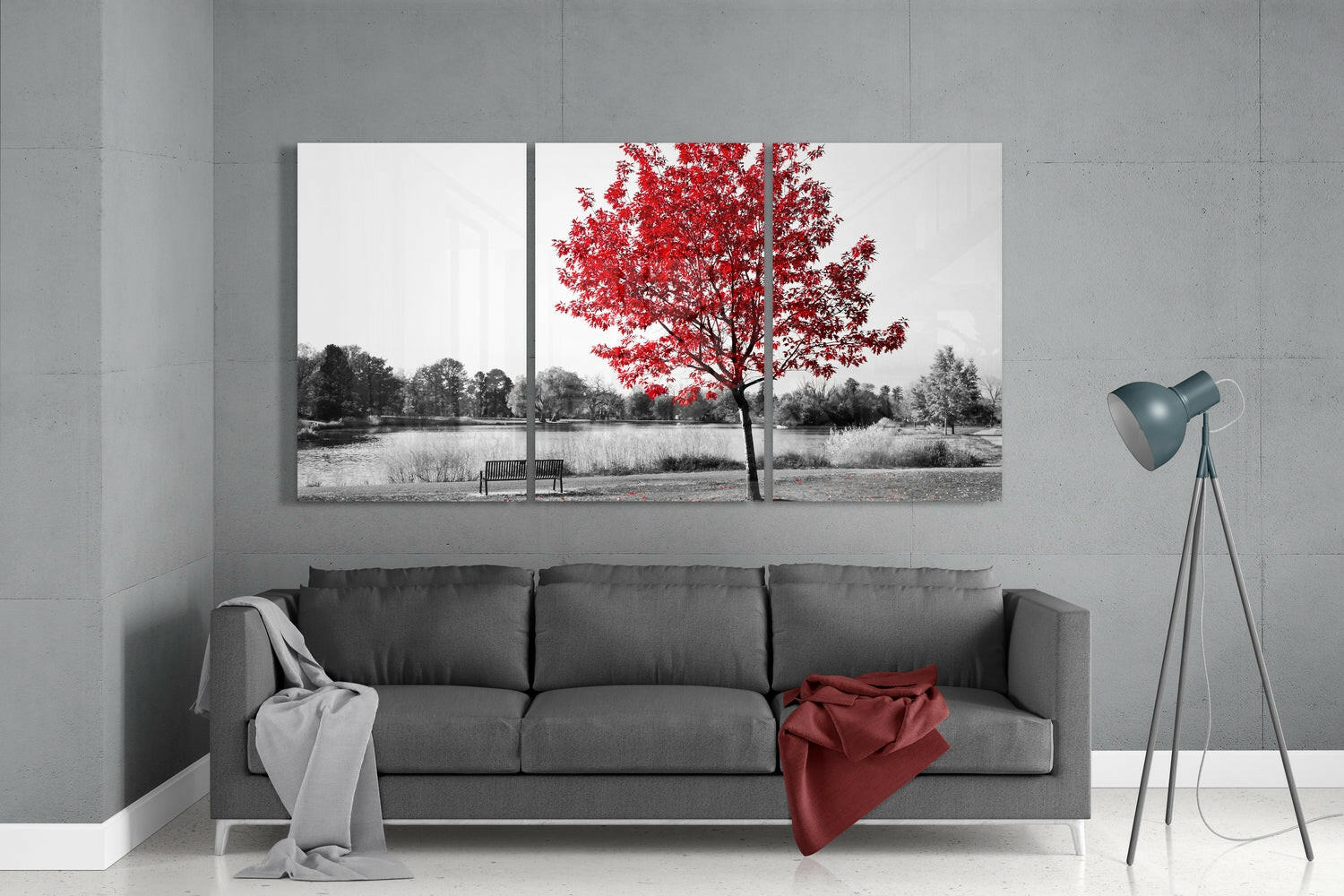 Three-panel glass wall art depicting a black and white landscape with a vibrant red tree, displayed above a gray sofa in a modern living room.