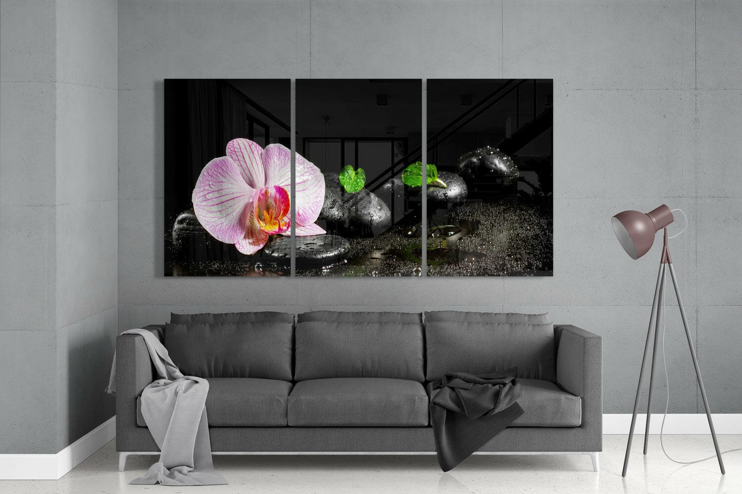Three-panel glass wall art displaying pink orchid flower and black stones with green foliage against grey living room wall above sofa.