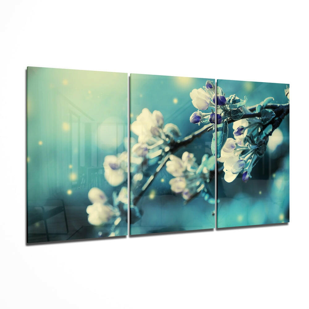 Three-panel glass wall art displaying delicate white and purple flowers on branches against a soft teal and yellow bokeh background.