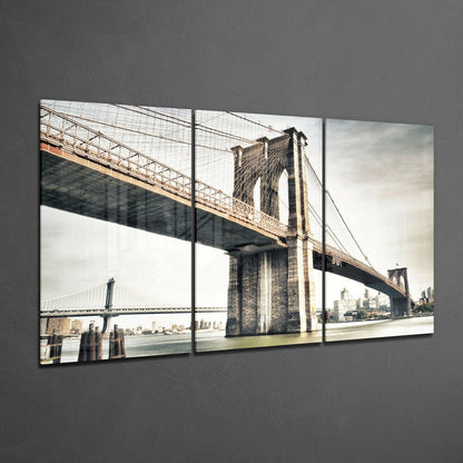 Three-panel glass wall art displaying the Brooklyn Bridge spanning the East River with Manhattan skyline in sepia tones against gray wall.