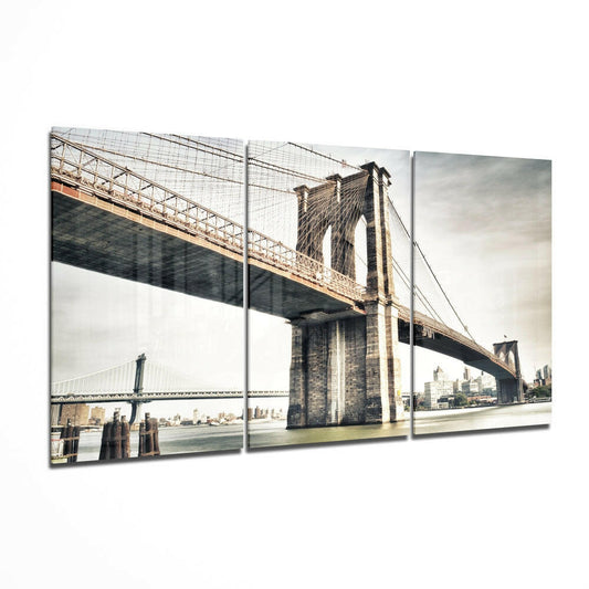 Three-panel glass wall art displaying the Brooklyn Bridge spanning the East River with Manhattan skyline in background under cloudy sky.