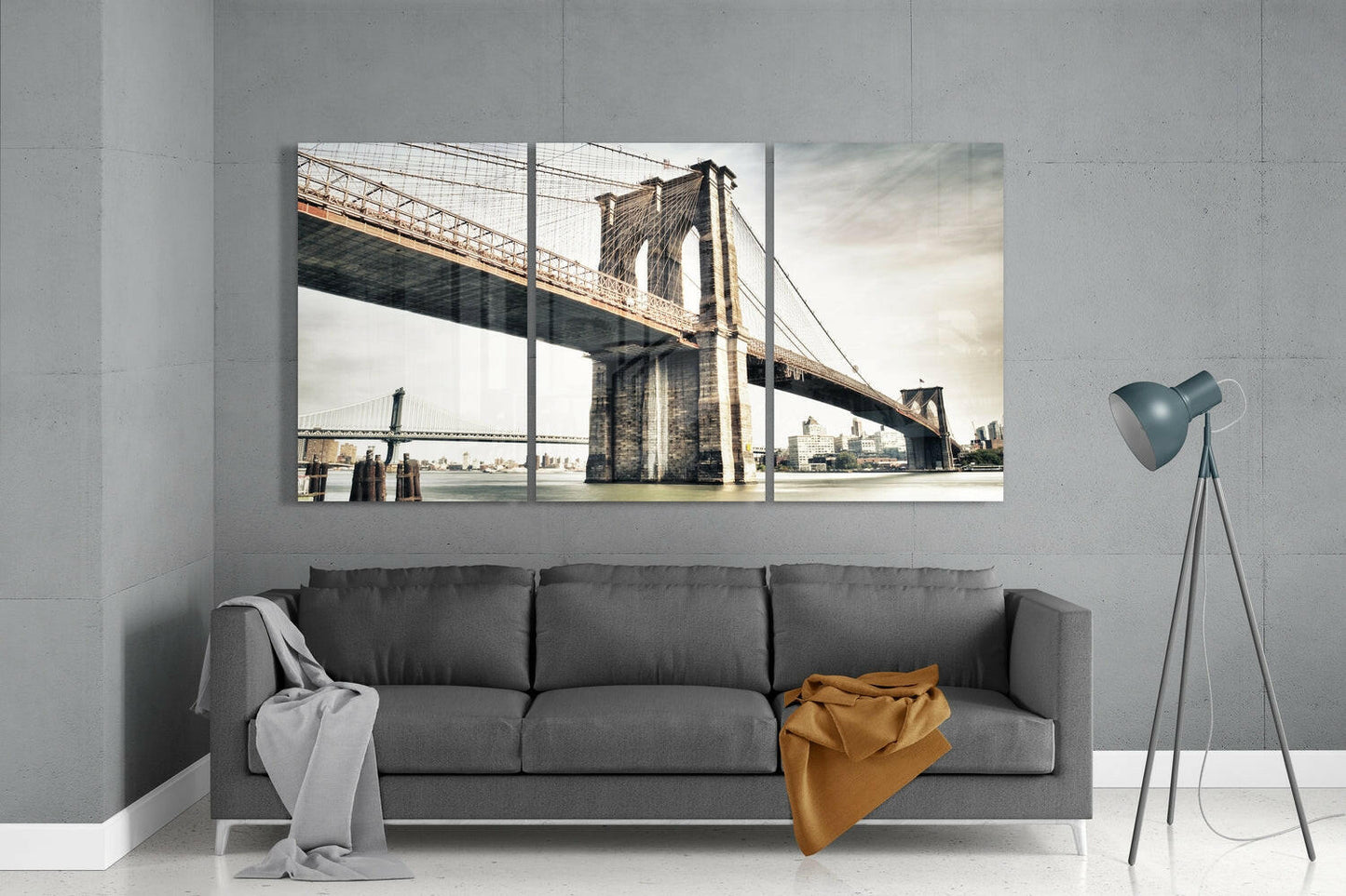 Three-panel glass wall art displaying Brooklyn Bridge with Manhattan skyline, mounted above gray sofa in minimalist interior.