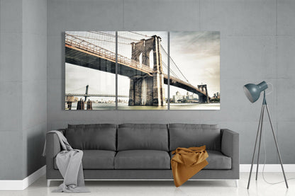 Three-panel glass wall art displaying Brooklyn Bridge with Manhattan skyline, mounted above gray sofa in minimalist interior.