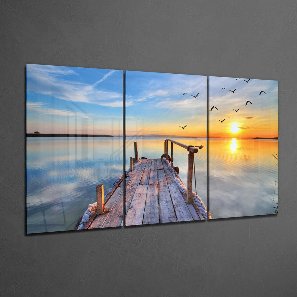 Three-panel glass wall art displaying a wooden dock extending into calm water at sunset, with flying birds and golden sky reflected in the water.