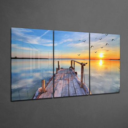 Three-panel glass wall art displaying a wooden dock extending into calm water at sunset, with flying birds and golden sky reflected in the water.