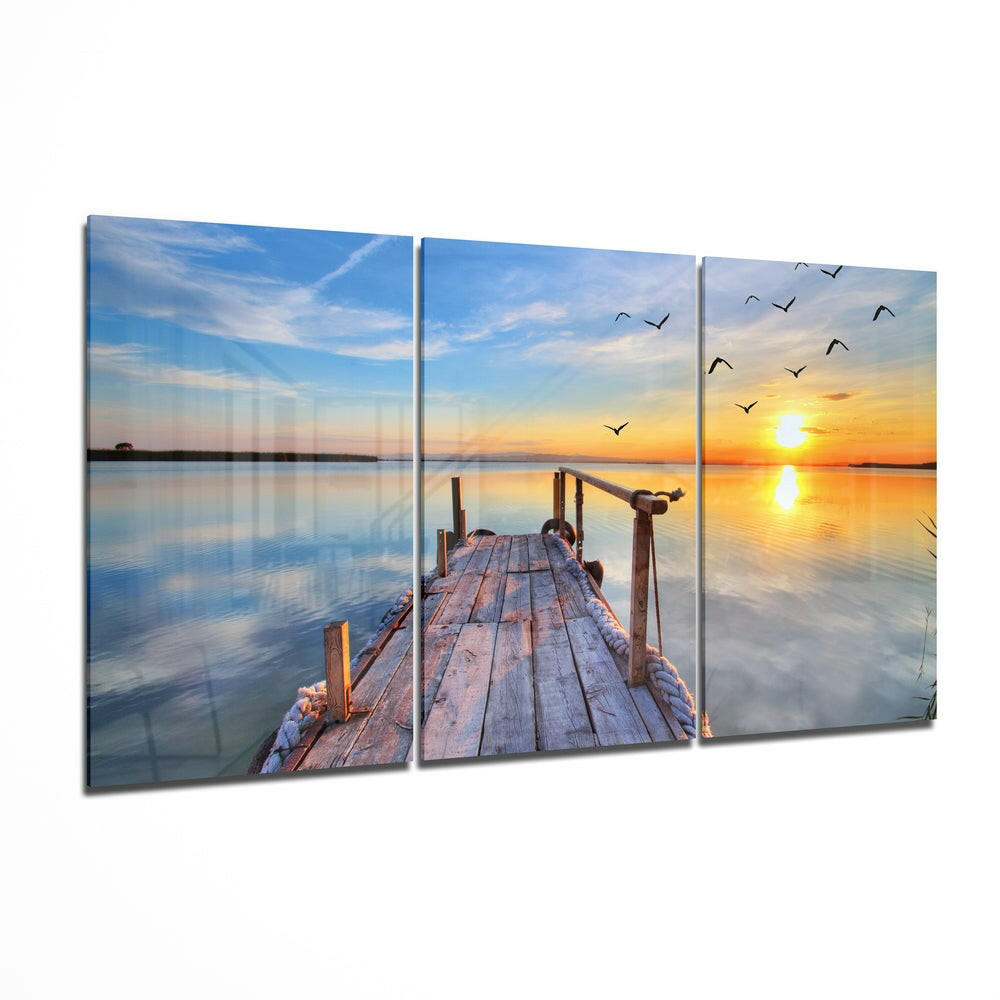 Three-panel glass wall art showing a wooden dock extending into calm water at sunset with birds flying overhead and golden sky reflection.