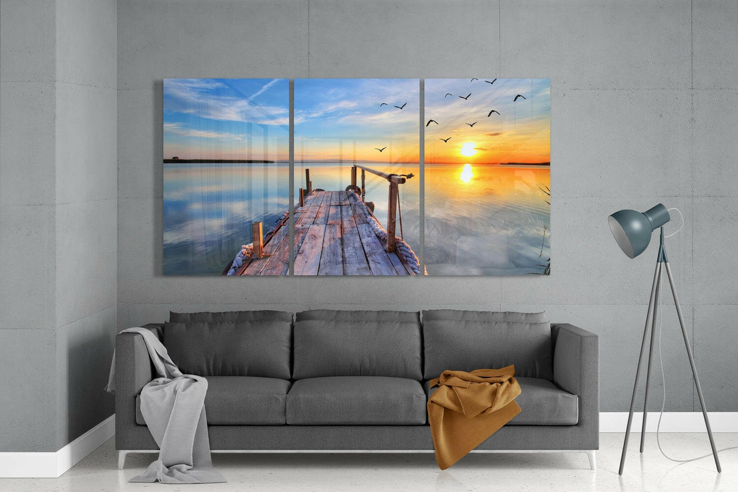Three-panel glass wall art displaying a wooden dock extending into calm water at sunset, with golden sky and flying birds, hung above a gray sofa in a modern living room.