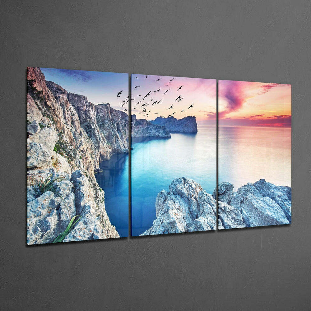 Three-panel glass wall art depicting rocky coastal cliffs, turquoise sea, and pink sunset sky with flying birds, mounted on gray wall.