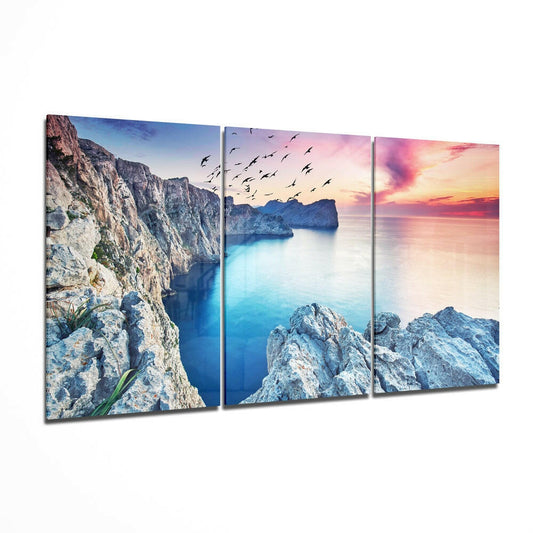 Three-panel glass wall art displaying rocky coastline cliffs, turquoise sea, and pink sunset sky with flying birds.