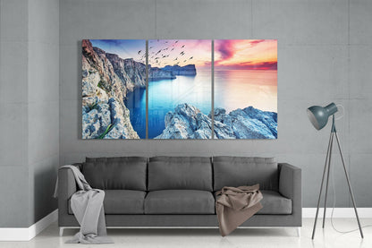 Three-panel glass wall art above gray sofa displaying rocky coastal cliffs, turquoise sea, and sunset sky with flying birds.