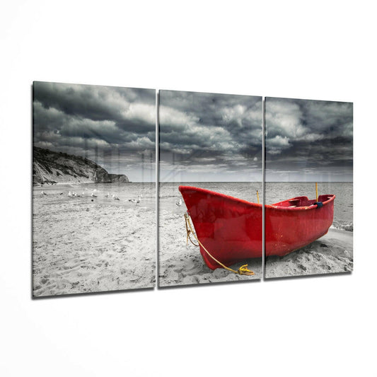Three-panel glass wall art showing a bright red rowboat beached on sand beneath stormy grey clouds.