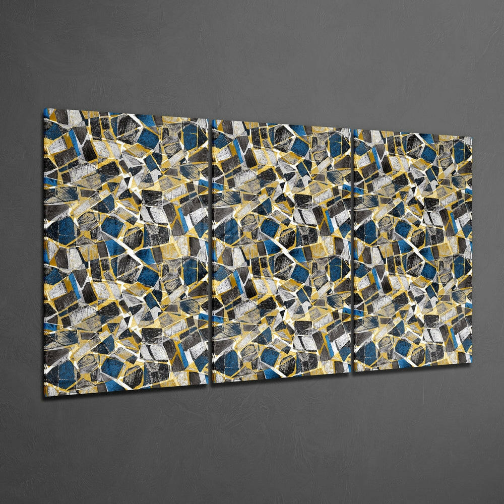 Three-panel glass wall art with abstract geometric pattern in navy blue, gold, white, and black shapes displayed on gray wall.