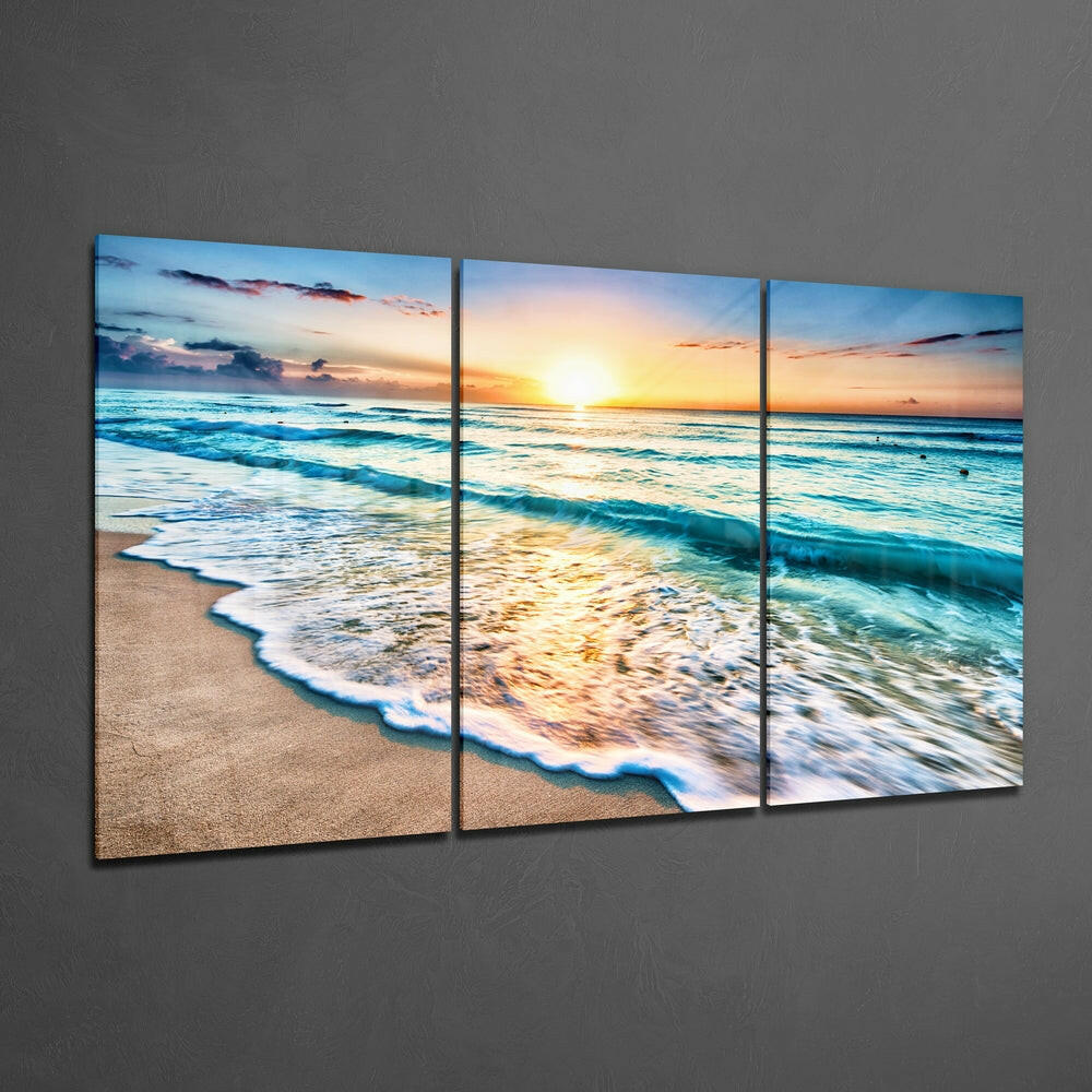 Three-panel glass wall art displaying a beach sunset with turquoise waves, golden light, and sandy shore mounted on gray wall.