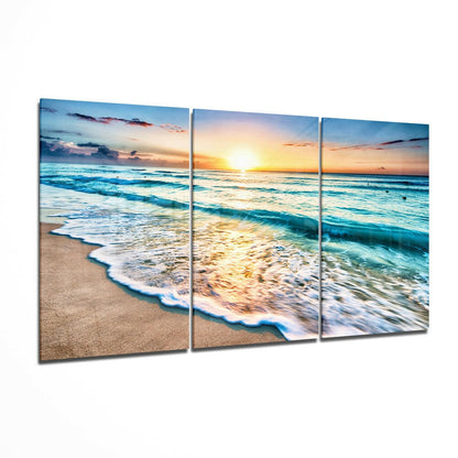 Three-panel glass wall art displaying a beach sunset with turquoise waves, golden light, and sandy shore in perspective view.