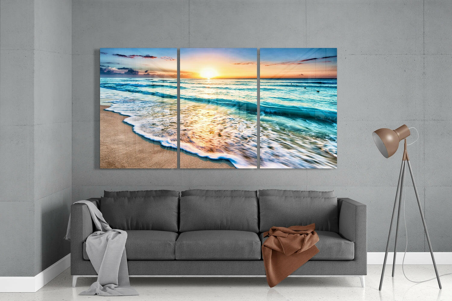 Three-panel triptych glass art depicting a sunset beach scene with turquoise waves and golden sand, displayed above a gray sofa in a modern living room.