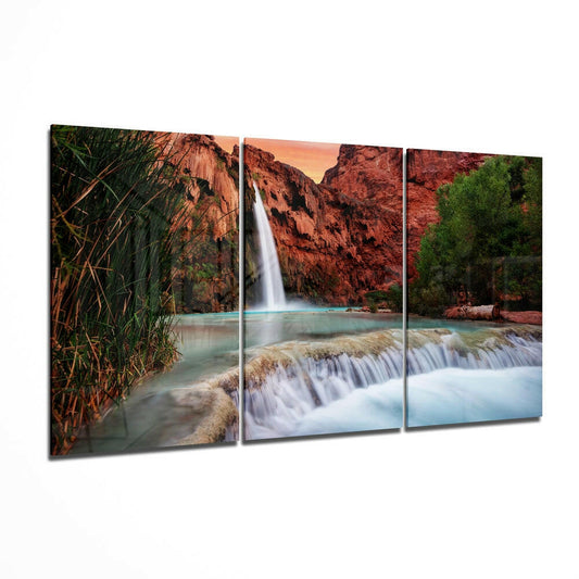 Three-panel glass wall art displaying a canyon waterfall with turquoise water, red rock cliffs, cascading streams, and green vegetation at sunset.