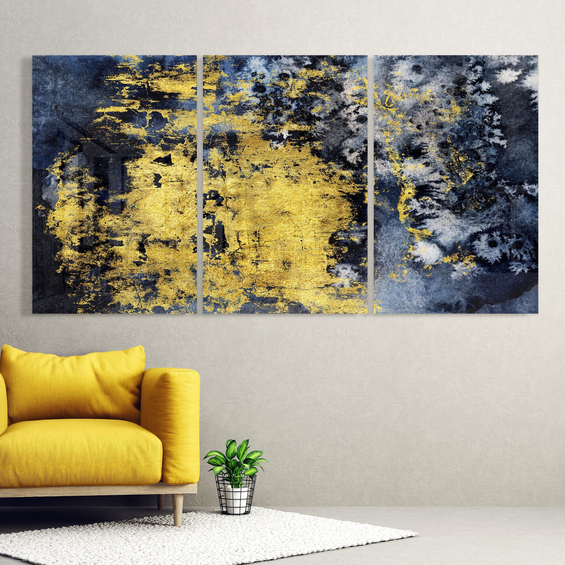 Three-panel triptych glass wall art with distressed yellow and gold paint splatters on dark blue background, displayed above yellow sofa.