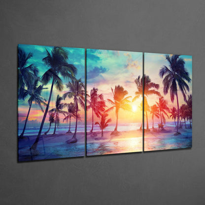 Three-panel glass wall art displaying a tropical beach sunset with palm trees, vibrant orange and pink sky, and blue ocean water.