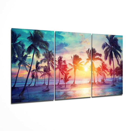 Three-panel glass wall art displaying a tropical beach sunset with palm trees, golden sun, and vibrant orange-pink-blue sky reflections.