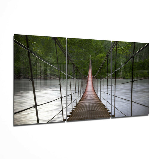 Three-panel glass wall art displaying a symmetrical perspective view of a red rope bridge stretching over a river through green forest.