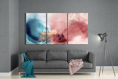 Three-panel glass wall art above gray sofa displaying blue and pink watercolor abstractions with gold accents.