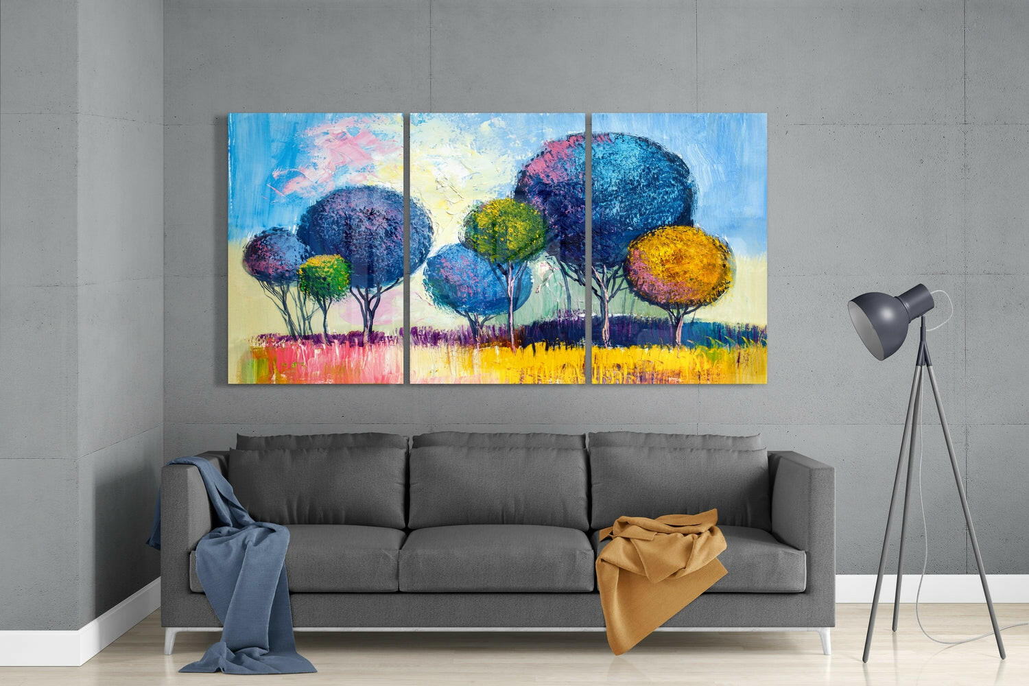 Colorful three-panel glass artwork depicting stylized trees with blue, yellow, and pink foliage above a yellow field, displayed above a gray sofa.