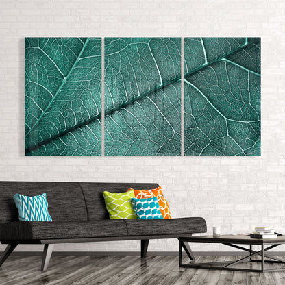 Three-panel green leaf vein glass art installed above a gray sofa with colorful patterned pillows in a modern living room.