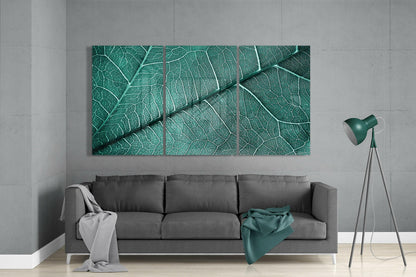 Three-panel green leaf vein glass art displayed above gray sofa in minimalist living room with teal lamp.