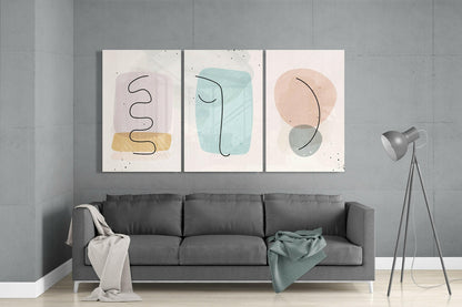 Three abstract glass wall art panels with pastel shapes and line drawings displayed above a gray sofa in a modern living room.