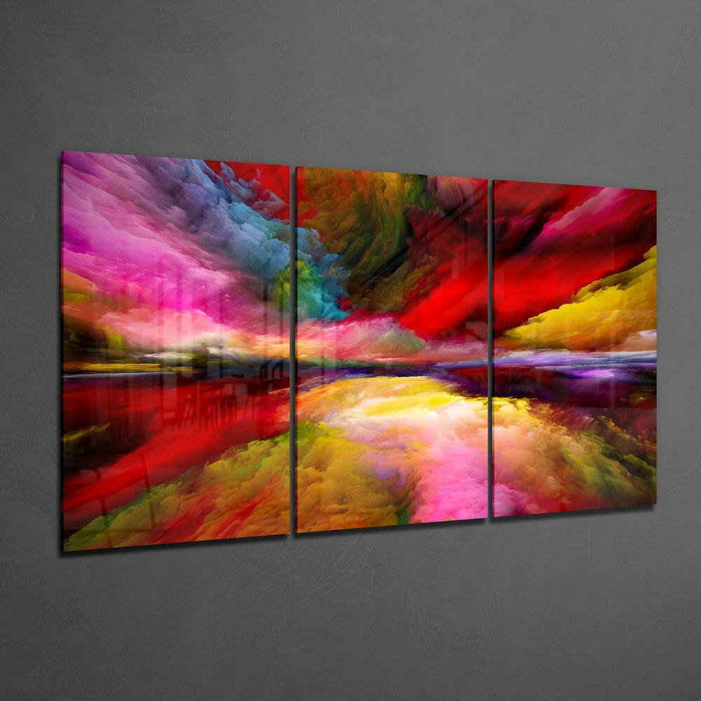 Three-panel glass wall art displaying a vibrant abstract landscape with swirling pink, red, yellow, and blue clouds reflected in water, mounted on gray wall.