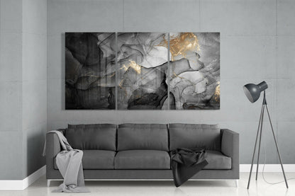 Three-panel abstract glass wall art with black, gray, and gold marbling hung above a gray sofa in a minimalist interior.