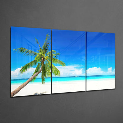 Three-panel glass wall art displaying a tropical beach scene with a palm tree leaning over turquoise water and white sand against a blue sky.