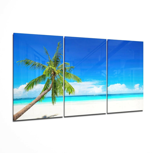 Three-panel glass wall art displaying a tropical beach scene with a leaning palm tree, white sand, turquoise water, and blue sky.