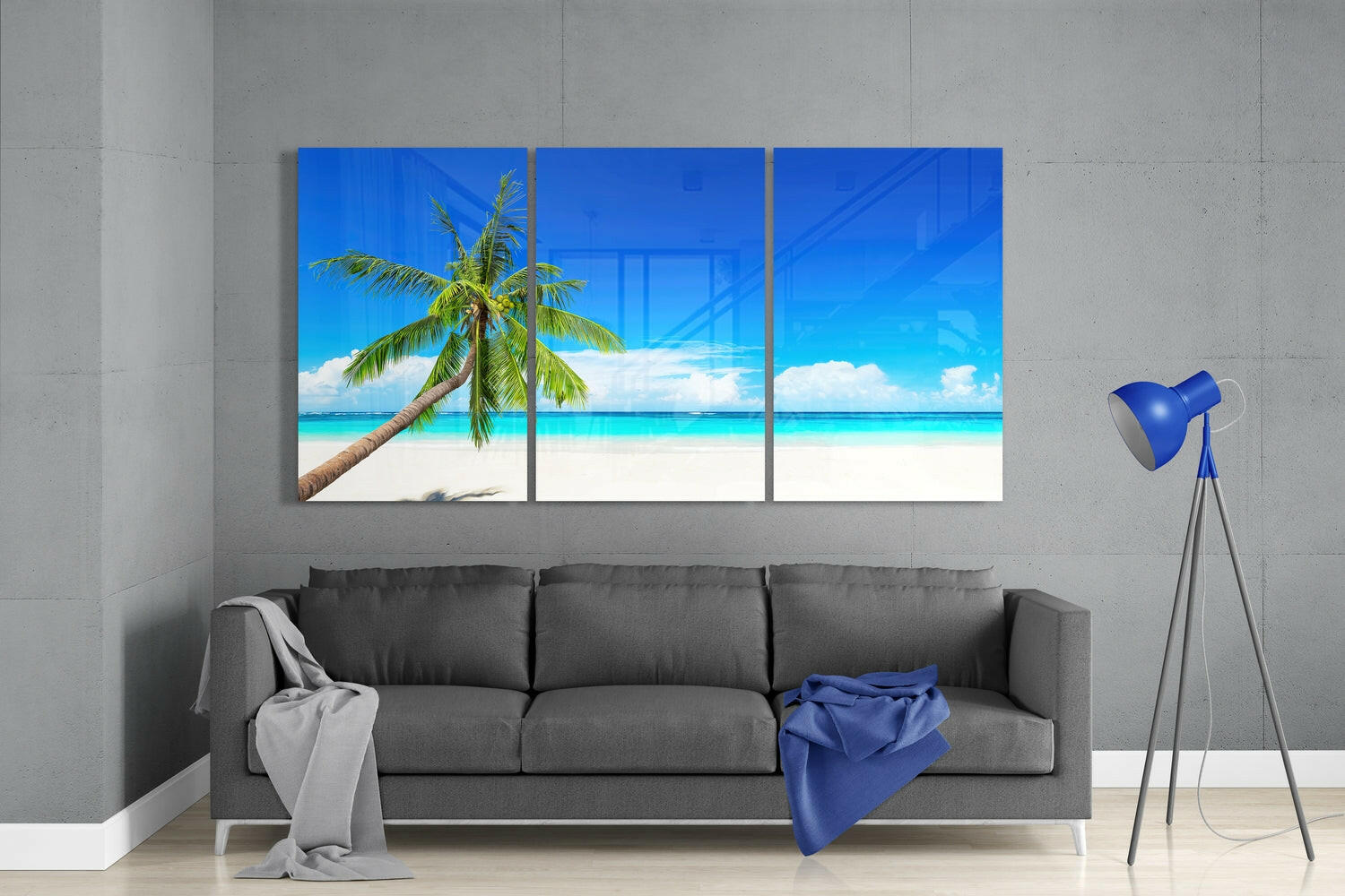 Three-panel blue glass beach artwork featuring a palm tree and turquoise ocean, displayed above a gray sofa in a modern interior.