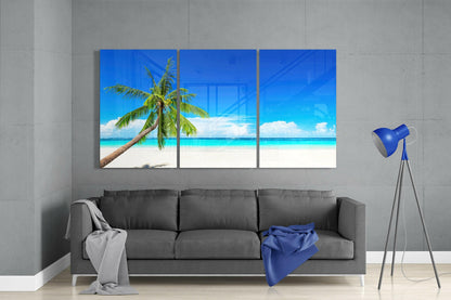 Three-panel blue glass beach artwork featuring a palm tree and turquoise ocean, displayed above a gray sofa in a modern interior.