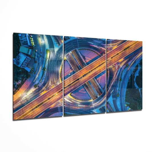 Three-panel glass wall art showing aerial nighttime view of illuminated highway interchanges with orange and blue light trails on dark roads.