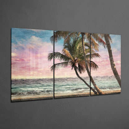 Three-panel glass wall art depicting a tropical beach sunset with palm trees leaning over turquoise water and pink-hued sky.