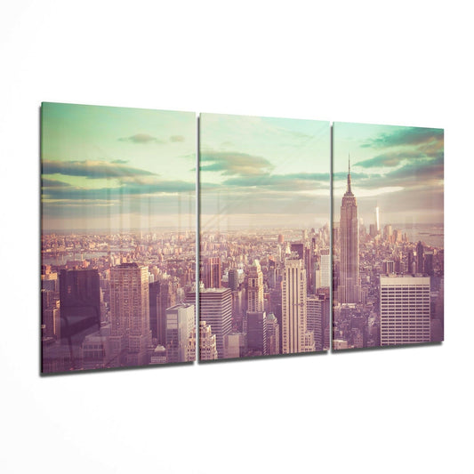 Three-panel glass wall art displaying a vintage-filtered Manhattan skyline with Empire State Building at sunset, soft pastel tones of pink, green, and gold.