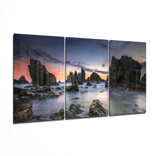 Three-panel glass wall art displaying rocky sea stacks at sunset with misty waves and colorful twilight sky.