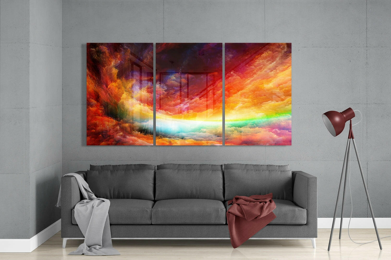 Three-panel glass wall art above gray sofa displaying vibrant red, orange, yellow, and rainbow-colored cloud landscape.