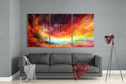 Three-panel glass wall art above gray sofa displaying vibrant red, orange, yellow, and rainbow-colored cloud landscape.